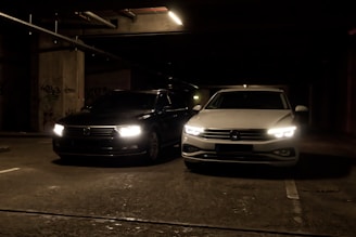 Two cars are parked side by side in a dimly lit parking garage. The headlights are turned on, illuminating the dark surroundings. The left car is black while the right car is white, and both are positioned near concrete pillars covered with graffiti.