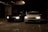 Two cars are parked side by side in a dimly lit parking garage. The headlights are turned on, illuminating the dark surroundings. The left car is black while the right car is white, and both are positioned near concrete pillars covered with graffiti.