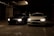 Two cars are parked side by side in a dimly lit parking garage. The headlights are turned on, illuminating the dark surroundings. The left car is black while the right car is white, and both are positioned near concrete pillars covered with graffiti.