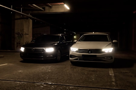 Two cars are parked side by side in a dimly lit parking garage. The headlights are turned on, illuminating the dark surroundings. The left car is black while the right car is white, and both are positioned near concrete pillars covered with graffiti.