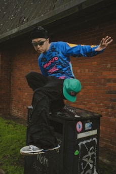 A person wearing a colorful blue and black outfit with sunglasses performs an athletic movement on a graffitied black metal structure. The background features a red brick wall and green grass.