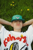 A person wearing a white t-shirt with bold red and black lettering and a teal cap lies on green grass. Yellow dandelions are scattered around, and one dandelion is placed on the shirt.