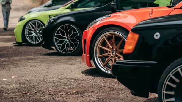 Several cars are parked in a line with visible wheels and fenders. The cars are of different colors including black, green, and orange. Each car has distinctive large alloy rims and low-profile tires, highlighting a sporty and modified appearance.