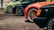 Several cars are parked in a line with visible wheels and fenders. The cars are of different colors including black, green, and orange. Each car has distinctive large alloy rims and low-profile tires, highlighting a sporty and modified appearance.