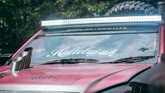 A close-up view of a customized car with a bright light bar mounted on the roof. The windshield displays the word 'Hallelujah' in stylized white lettering. The vehicle is painted in a matte red color. Large off-road tires are visible in the background, along with a portion of a metallic exhaust pipe protruding from the hood. Trees are reflected in the windshield.