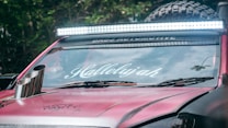 A close-up view of a customized car with a bright light bar mounted on the roof. The windshield displays the word 'Hallelujah' in stylized white lettering. The vehicle is painted in a matte red color. Large off-road tires are visible in the background, along with a portion of a metallic exhaust pipe protruding from the hood. Trees are reflected in the windshield.