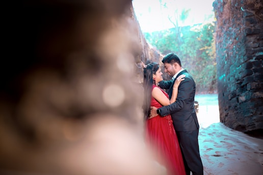 A couple is embracing lovingly in an outdoor setting with stone walls and soft sand underfoot. The woman is wearing a vibrant red dress, while the man is dressed in a formal black suit. The background is blurred, featuring green foliage and natural light.