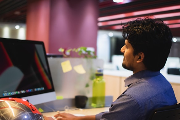 A person in a blue shirt sits at a desk in an office setting, looking at a large computer monitor. The desk has various items, including a green water bottle, sticky notes on a partition, and a decorative object resembling a helmet. The background features a modern office interior with muted lighting and a plant visible in the background.