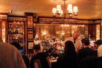 A warmly lit bar scene with a mirror reflecting bottles lined up on the counter. A bartender in a white shirt and black tie serves customers who are seated at the bar. The walls are decorated with artistic silhouettes and framed pictures. Several patrons are engaging in conversation, creating a lively yet cozy atmosphere.