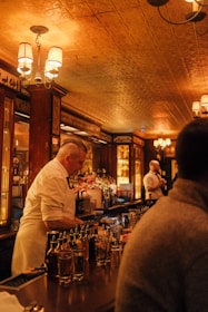 A warmly lit bar scene featuring a bartender serving drinks. The bar is stocked with various bottles, and another person stands nearby. The atmosphere is cozy with a vintage-style ceiling, ornate lighting fixtures, and wooden decor.