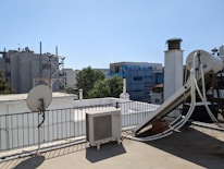 A rooftop scene featuring various equipment including a satellite dish, an outdoor air conditioning unit, and a large cylindrical tank with pipes connected to it. The roof is bordered by a metal railing and some structures can be seen in the background, including buildings covered in scaffolding and some trees.