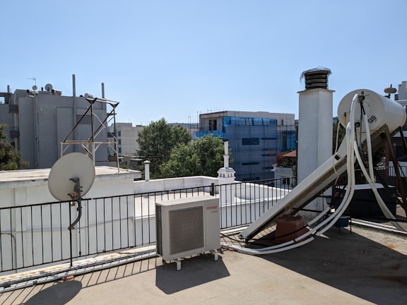 A rooftop scene featuring various equipment including a satellite dish, an outdoor air conditioning unit, and a large cylindrical tank with pipes connected to it. The roof is bordered by a metal railing and some structures can be seen in the background, including buildings covered in scaffolding and some trees.