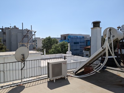 A rooftop scene featuring various equipment including a satellite dish, an outdoor air conditioning unit, and a large cylindrical tank with pipes connected to it. The roof is bordered by a metal railing and some structures can be seen in the background, including buildings covered in scaffolding and some trees.