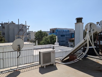 A rooftop scene featuring various equipment including a satellite dish, an outdoor air conditioning unit, and a large cylindrical tank with pipes connected to it. The roof is bordered by a metal railing and some structures can be seen in the background, including buildings covered in scaffolding and some trees.