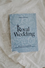 A book titled 'Royal Wedding' by Marina Minelli rests on an ornate white lace fabric. The book has a light blue cover with elegant floral patterns and bold black text. At the bottom, there is a subtitle in Italian.