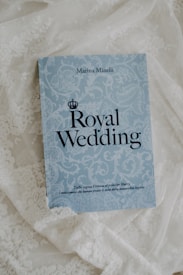 A book titled 'Royal Wedding' by Marina Minelli rests on an ornate white lace fabric. The book has a light blue cover with elegant floral patterns and bold black text. At the bottom, there is a subtitle in Italian.