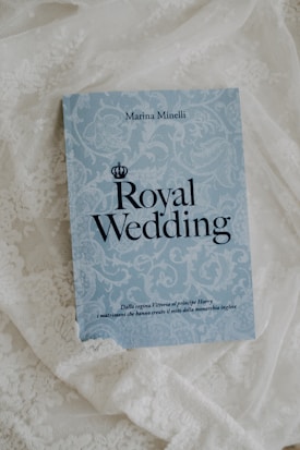A book titled 'Royal Wedding' by Marina Minelli rests on an ornate white lace fabric. The book has a light blue cover with elegant floral patterns and bold black text. At the bottom, there is a subtitle in Italian.