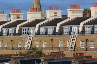 A row of brick townhouses with multiple chimneys, white framed windows, and rooftop terraces. The buildings feature steep slate roofs and external stairways. A church steeple rises in the background, while a clear blue sky provides a serene backdrop.