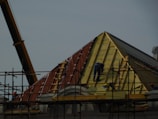 A construction site featuring workers on scaffolding constructing a steep, triangular roof structure. The workers are clad in safety gear, and the structure is partially covered with insulation and roofing materials. A crane is visible on the left side contributing to the construction effort. The overall setting suggests an active work environment focused on building or repairing the roof.