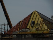 A construction site featuring workers on scaffolding constructing a steep, triangular roof structure. The workers are clad in safety gear, and the structure is partially covered with insulation and roofing materials. A crane is visible on the left side contributing to the construction effort. The overall setting suggests an active work environment focused on building or repairing the roof.