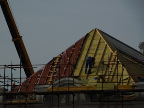 A construction site featuring workers on scaffolding constructing a steep, triangular roof structure. The workers are clad in safety gear, and the structure is partially covered with insulation and roofing materials. A crane is visible on the left side contributing to the construction effort. The overall setting suggests an active work environment focused on building or repairing the roof.