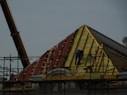 A construction site featuring workers on scaffolding constructing a steep, triangular roof structure. The workers are clad in safety gear, and the structure is partially covered with insulation and roofing materials. A crane is visible on the left side contributing to the construction effort. The overall setting suggests an active work environment focused on building or repairing the roof.