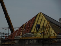 A construction site featuring workers on scaffolding constructing a steep, triangular roof structure. The workers are clad in safety gear, and the structure is partially covered with insulation and roofing materials. A crane is visible on the left side contributing to the construction effort. The overall setting suggests an active work environment focused on building or repairing the roof.