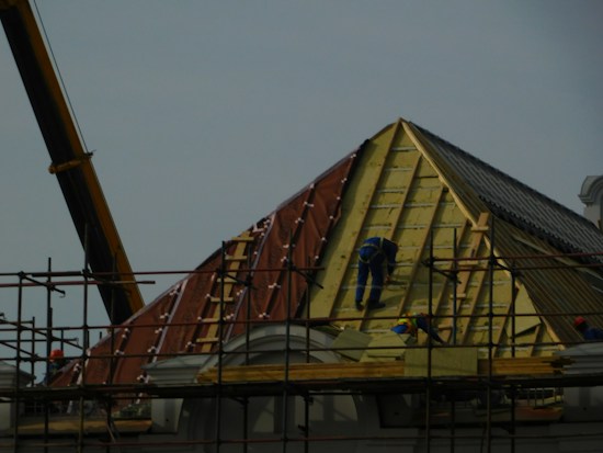 A construction site featuring workers on scaffolding constructing a steep, triangular roof structure. The workers are clad in safety gear, and the structure is partially covered with insulation and roofing materials. A crane is visible on the left side contributing to the construction effort. The overall setting suggests an active work environment focused on building or repairing the roof.