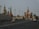 A cityscape featuring iconic Russian architecture, with colorful onion domes and intricately designed towers. A large road with multiple lanes stretches across the foreground, occupied by several cars and a few pedestrians.