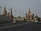 A cityscape featuring iconic Russian architecture, with colorful onion domes and intricately designed towers. A large road with multiple lanes stretches across the foreground, occupied by several cars and a few pedestrians.