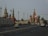 A cityscape featuring iconic Russian architecture, with colorful onion domes and intricately designed towers. A large road with multiple lanes stretches across the foreground, occupied by several cars and a few pedestrians.