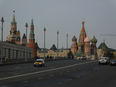 A cityscape featuring iconic Russian architecture, with colorful onion domes and intricately designed towers. A large road with multiple lanes stretches across the foreground, occupied by several cars and a few pedestrians.