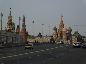 A cityscape featuring iconic Russian architecture, with colorful onion domes and intricately designed towers. A large road with multiple lanes stretches across the foreground, occupied by several cars and a few pedestrians.