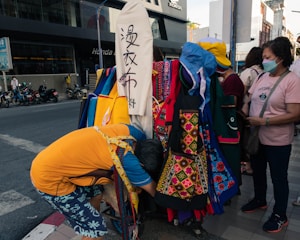 A street scene with people gathered around a stall displaying colorful textile products, including aprons with intricate patterns. The people appear to be examining or shopping for these items. Motorcycles are parked on the street in the background, and a person is bending over, possibly arranging or searching through the textile goods.
