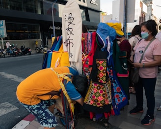 A street scene with people gathered around a stall displaying colorful textile products, including aprons with intricate patterns. The people appear to be examining or shopping for these items. Motorcycles are parked on the street in the background, and a person is bending over, possibly arranging or searching through the textile goods.