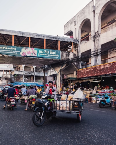 A bustling street market scene with several people on motorbikes carrying goods. The market is under a large concrete structure with a bridge overhead displaying signs in a non-English language. Stalls with various items are visible in the background, and the market appears lively and busy.
