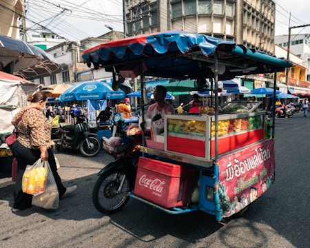 A bustling street scene featuring a mobile food cart filled with a variety of fresh fruits. The cart is covered with a colorful canopy and is situated on a busy street with people and motorbikes in the background. A woman is walking past carrying grocery bags while the vendor tends to the cart.