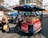 A bustling street scene featuring a mobile food cart filled with a variety of fresh fruits. The cart is covered with a colorful canopy and is situated on a busy street with people and motorbikes in the background. A woman is walking past carrying grocery bags while the vendor tends to the cart.