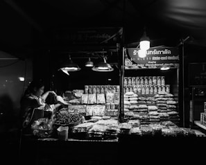 A vendor is preparing goods at a market stall that displays an array of packaged and loose dry goods, predominantly nuts and grains. The stall is illuminated by several hanging lights, casting a bright glow over the products. The background signage includes both text and graphics, indicating the type of goods being sold.