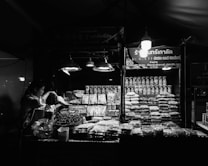 A vendor is preparing goods at a market stall that displays an array of packaged and loose dry goods, predominantly nuts and grains. The stall is illuminated by several hanging lights, casting a bright glow over the products. The background signage includes both text and graphics, indicating the type of goods being sold.