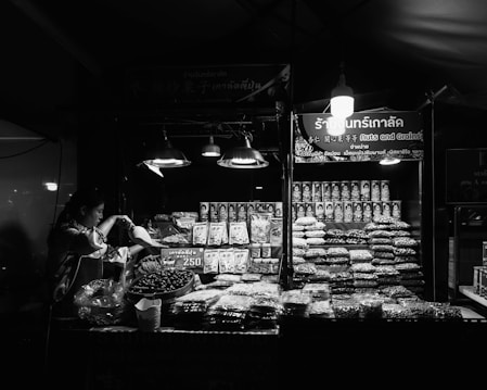 A vendor is preparing goods at a market stall that displays an array of packaged and loose dry goods, predominantly nuts and grains. The stall is illuminated by several hanging lights, casting a bright glow over the products. The background signage includes both text and graphics, indicating the type of goods being sold.