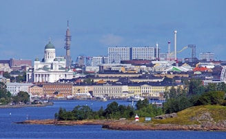 A scenic view of a cityscape featuring a prominent cathedral with a green dome, surrounded by various buildings. In the background, there are modern structures with game rides, possibly part of an amusement park, and a tower. The foreground includes a body of water with small islands covered in vegetation.