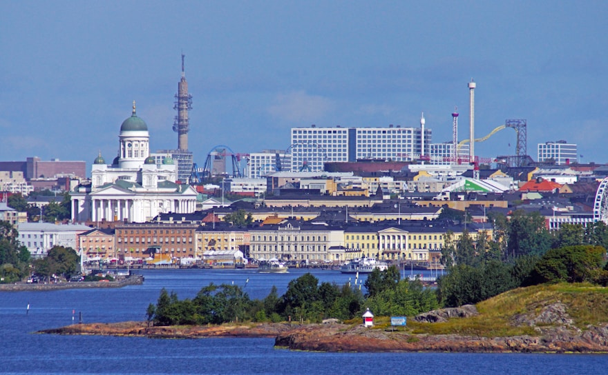 A scenic view of a cityscape featuring a prominent cathedral with a green dome, surrounded by various buildings. In the background, there are modern structures with game rides, possibly part of an amusement park, and a tower. The foreground includes a body of water with small islands covered in vegetation.