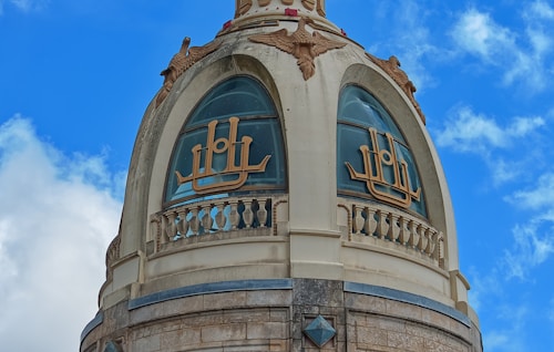 An ornate dome structure featuring intricate carvings and designs with golden script on the glass. The architecture includes detailed embellishments and sculptural elements, set against a bright blue sky with scattered clouds.