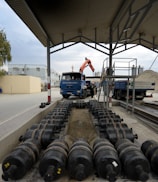 A parking area with a blue truck under a metal canopy. Several large cylindrical weights are aligned on the ground, and an excavator is visible in the background next to a mound of sand. Industrial buildings and trees are in the distance.