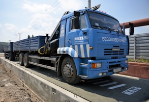 A large blue truck is parked on a weighing platform. The truck has multiple wheels and a flatbed trailer. It features text and logos on the side, and there is a metal fence in the background along with a clear sky.