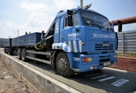 A large blue truck is parked on a weighing platform. The truck has multiple wheels and a flatbed trailer. It features text and logos on the side, and there is a metal fence in the background along with a clear sky.
