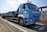 A large blue truck is parked on a weighing platform. The truck has multiple wheels and a flatbed trailer. It features text and logos on the side, and there is a metal fence in the background along with a clear sky.