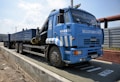 A large blue truck is parked on a weighing platform. The truck has multiple wheels and a flatbed trailer. It features text and logos on the side, and there is a metal fence in the background along with a clear sky.