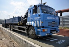 A large blue truck is parked on a weighing platform. The truck has multiple wheels and a flatbed trailer. It features text and logos on the side, and there is a metal fence in the background along with a clear sky.
